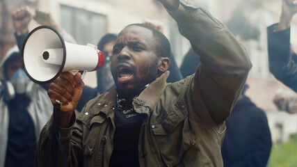 African-American young handsome man screaming in megaphone at protest for human rights outdoors in smoke. Group of people protesting at street. Strike against violence. - obrazy, fototapety, plakaty