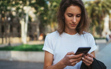 pretty woman reads  text message on mobile smartphone while standing in a Park on warm summer day, gorgeous woman listens to music on earphones and searches for information on cell phone outdoors