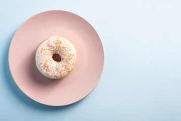 Vanilla donut with sprinkles on pink plate, sweet glazed dessert food on blue minimal background, top view copy space