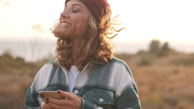 Happy blonde woman wearing hat and plaid shirt using smartphone and looking around outdoors