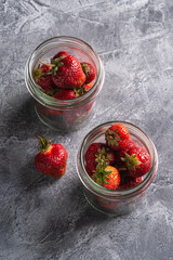 Fresh ripe strawberry fruits in two glass jars, summer vitamin berries on grey stone background, angle view