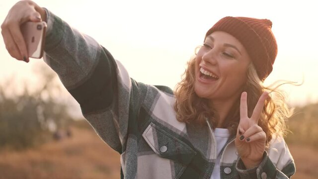 Happy blonde woman wearing hat and plaid shirt making selfie on smartphone outdoors