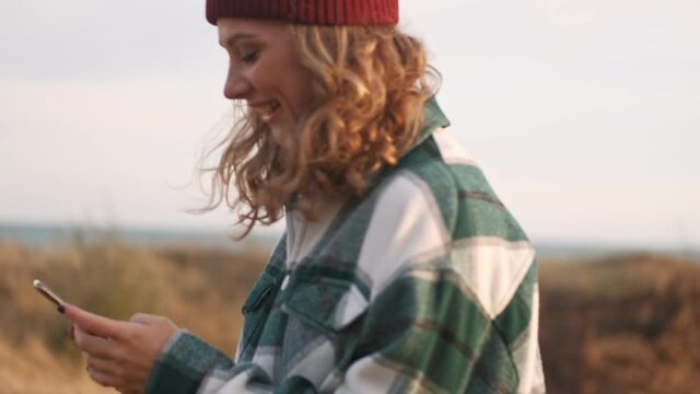 Side view of Happy blonde woman wearing hat and plaid shirt using smartphone while walking outdoors