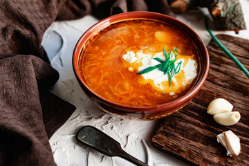 A portion of red borscht in a brown plate, next to an old wooden Board garlic and quail egg, green leeks and driftwood. White sour cream in the soup, next to an old Polish spoon