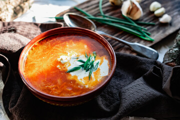 A portion of red borscht in a brown plate, next to an old wooden Board garlic and quail egg, green leeks and driftwood. White sour cream in the soup, next to an old Polish spoon
