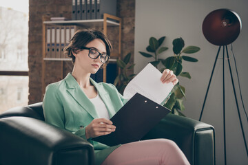 Concentrated minded leader marketer chief financier girl sit leather armchair read clip board analyze startup development progress presentation wear green blazer jacket in wokrstation