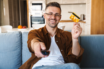 Image of handsome smiling man holding credit card while watching tv