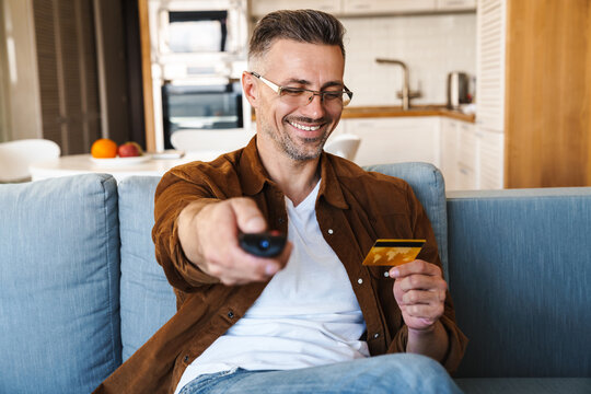 Image of handsome smiling man holding credit card while watching tv