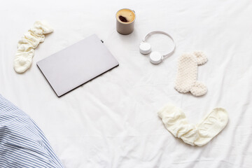 Weekend morning,  laptop, warm socks, cup of coffee, wireless headphones, mask for sleeping on bed sheet. Concept of relax at home, top view, flat lay with copy space.