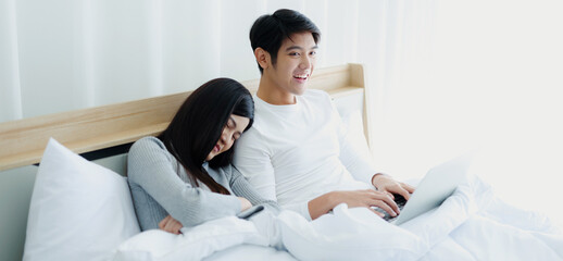 Obraz na płótnie Canvas The concept of happy and smiling Asian couples working at home on their laptops in bed.