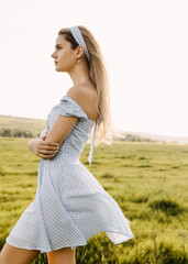 Fototapeta na wymiar Young beautiful woman with long blonde hair, in sun light, backlit, outdoors, wearing a light blue dress, standing in a field.