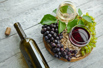 A cluster of dark and green grapes and two wine glasses with red and white wine, bottle on a round straw tray and grey wooden table. Wine composition flat lay.