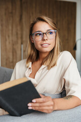 Image of young pleased woman reading book while sitting on couch