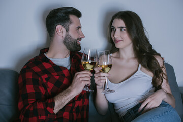 happy young couple sitting on sofa, looking at each other and clinking glasses of white wine