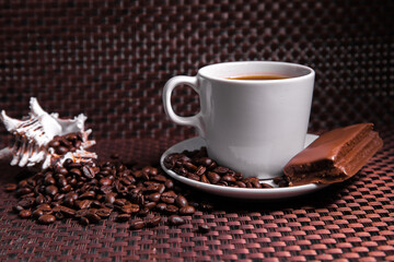 A cup of coffee with a chocolate bar, scattered coffee beans and a shell.