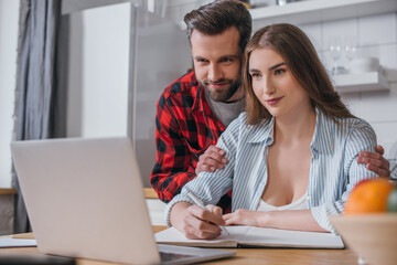 selective focus of smiling man touching shoulders of pretty girlfriend working at laptop