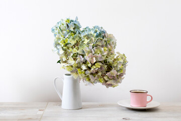 A large branch with green and blue pastel hydrangea in a white jug on a beige table.