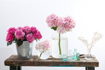 bouquet of red roses in a tin bucket, multicolored pastel colored hydrangeas in a glass jar, gypsophila in a flask and a set of small bottles on the old surface of a wooden bench
