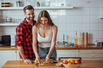 happy man looking at camera near attractive girlfriend preparing breakfast with fresh fruits