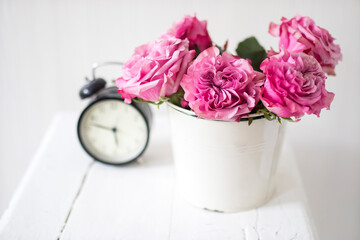 A bouquet of pink roses in a tin white bucket on an old stool with an alarm clock and a napkin with embroidery near the bed on a beige background