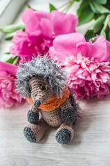 Crocheted hedgehog with pink peony flower on wooden background.