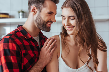 smiling man touching shoulder of happy young girlfriend in kitchen