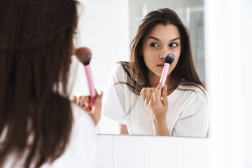 Photo of pleased woman using powder brush while looking at mirror