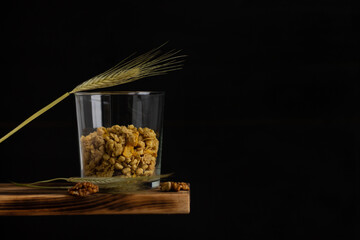 Cereal granola in a transparent glass with rye spike and walnuts on a shelf on a black background....