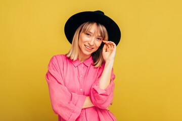 Young stylish trendy woman posing on camera. Beautiful cheerful girl hold one hand close to face and smile. Female model wear stylish pink shirt and black hat. Isolated over yellow background.