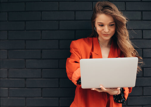 Confident busy young woman isolated over dark background. Businesswoman typing on laptop's keyboard. Remote work outside of office. Concentrated girl in orange jacket on picture.