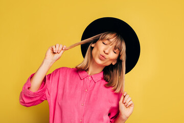 Picture of cheerful nice positive young woman posing on camera alone in studio. Girl in pink shirt and black hat hold her blonde hair in hands and keep eyes closed. Isolated over yellow background.