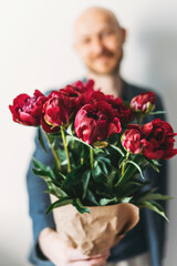 Attractive adult bearded man holding beautiful bouquet of dark peonies in craft paper on the grey background