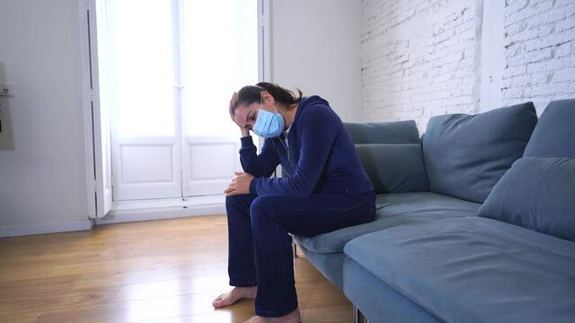 Sad latin woman with protective face mask at home living room couch feeling tired and worried suffering depression amid coronavirus lockdown and social distancing. Mental Health and isolation concept.