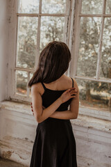 A girl stands in a black dress near the window and holds her hands behind her back.