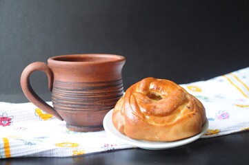 A clay mug and a roll stand on a white kitchen towel with flowers on a black background.