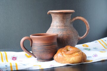 A clay jug, a mug and a roll stand on a white kitchen towel with flowers on a black background. 