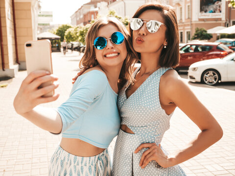 Two young beautiful smiling hipster girls in trendy summer clothes.Sexy carefree women posing on the street background in sunglasses. They taking selfie self portrait photos on smartphone at sunset