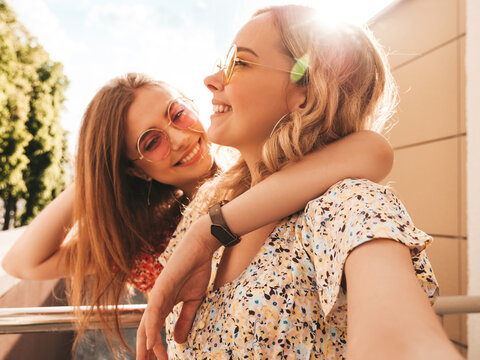 Two young beautiful smiling hipster girls in trendy summer sundress.Sexy carefree women posing on the street background in sunglasses. They taking selfie self portrait photos on smartphone at sunset