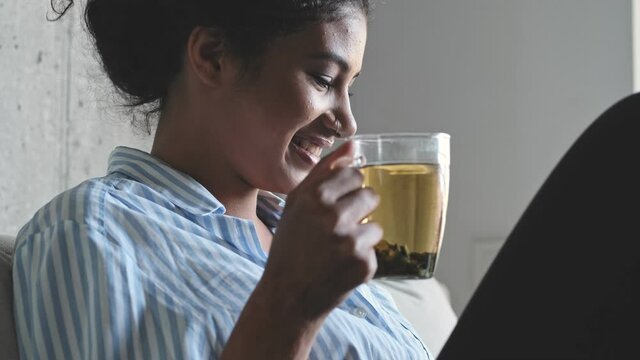 A happy glad young african woman is drinking tea and sitting on the sofa at home