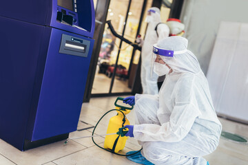 Professional workers in hazmat suits disinfecting ATM machine.