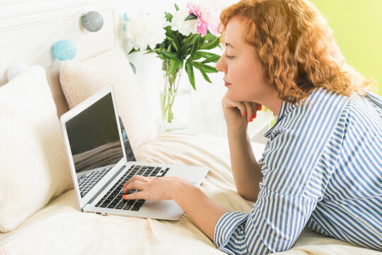 Woman with red hair at home with a laptop on the bed. Remote work and free time housewives. Surfing the internet