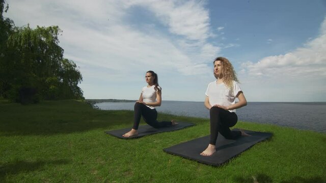 Two young women practice yoga on coast near the lake or sea. Training Crescent Lunge on the Knee pose.