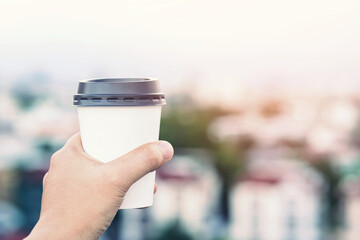 hand holding paper cup of take away drinking coffee on city background natural morning sunlight. space Place for your text or logo.