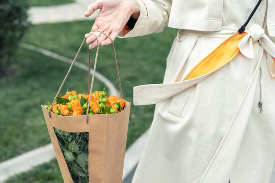 Close-up young adult fashion classy woman model hand holding authentic fresh orange spray roses flower bouquet wrapped in craft paper bag on city urban street outdoor. Spring present gift dilevery