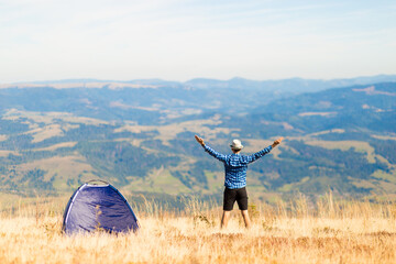 Young tourist man admire beautiful mountains view with arms up near tent