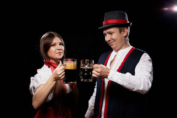 Happy Oktoberfest couple in national ethnic dress clinking beer mugs and black background. Girl and boy posing in studio with alcohol during celebration Oktoberfest.