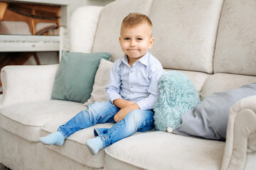 A beautiful blond boy smiles and poses. A sweet child, a happy childhood. sitting on a light sofa in a bright interior