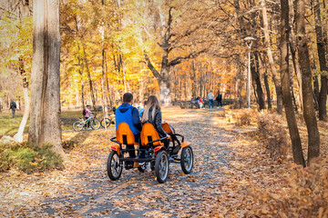 Couple people enjoy riding pedal surrey cart bike rent or sharing outdoors in beautiful golden autumn city park, garden or forest on bright warm day. Healthy family outside sport recreation activity
