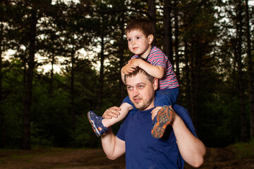 Dad carries his son on his shoulders around his neck. Father and son walk in the woods in the wild among the trees in the evening. Love and family