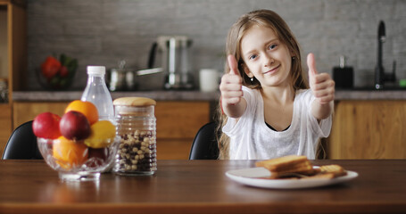 Beautiful schoolgirl with long hair sitting at the table in the kitchen. A cute child shows a thumb up or finger good.